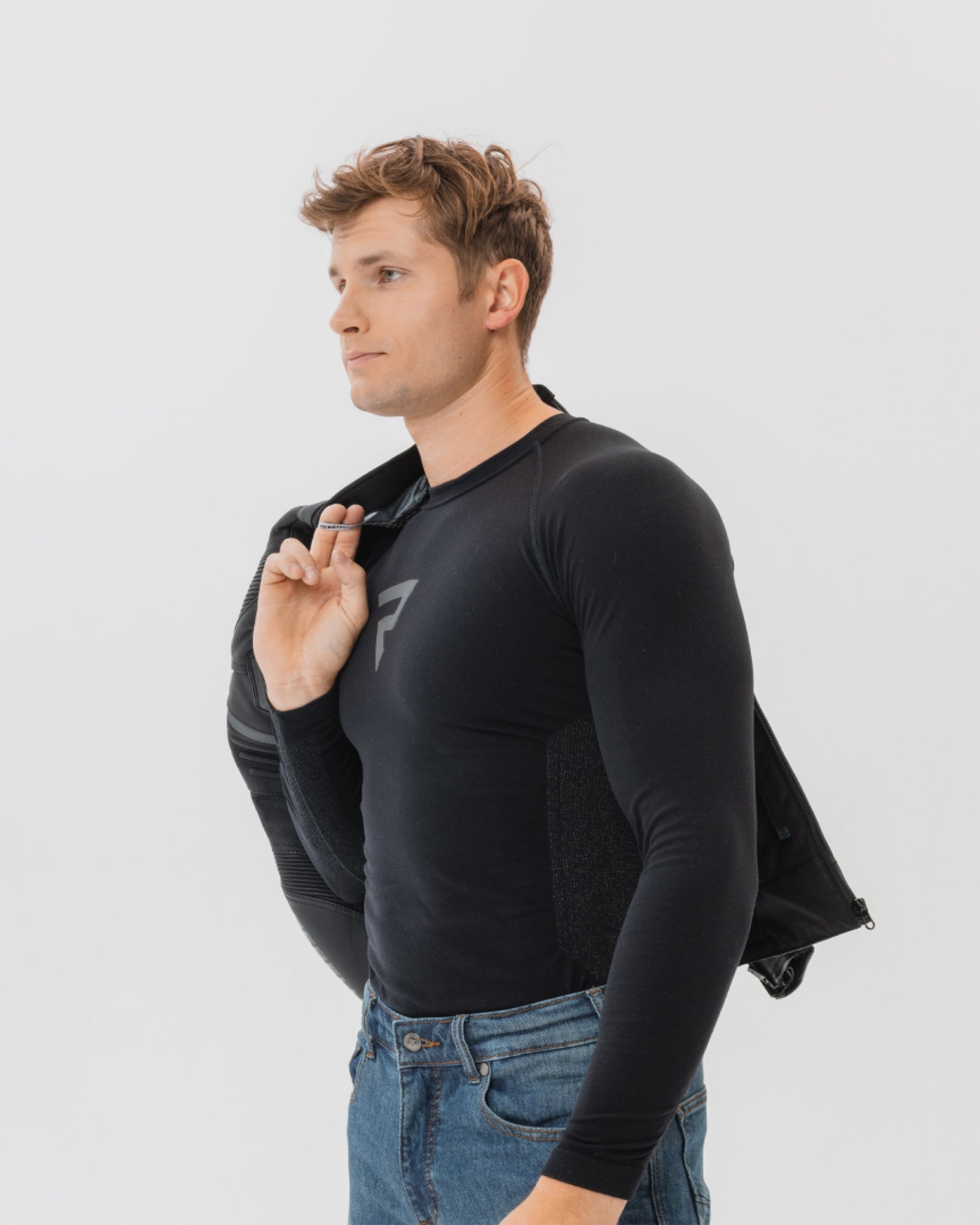 Man wearing a black long-sleeve shirt with a logo and blue jeans on a light gray background