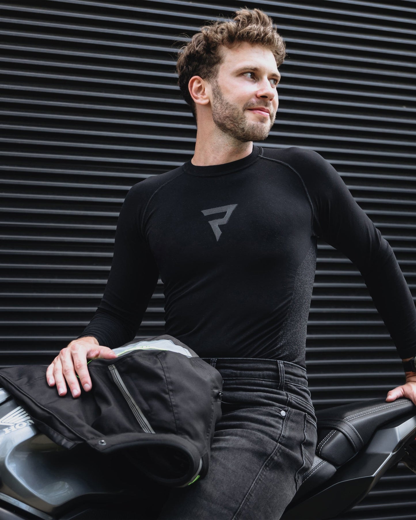 Man wearing a black long-sleeve shirt with a logo, sitting on a car against a black corrugated metal wall.